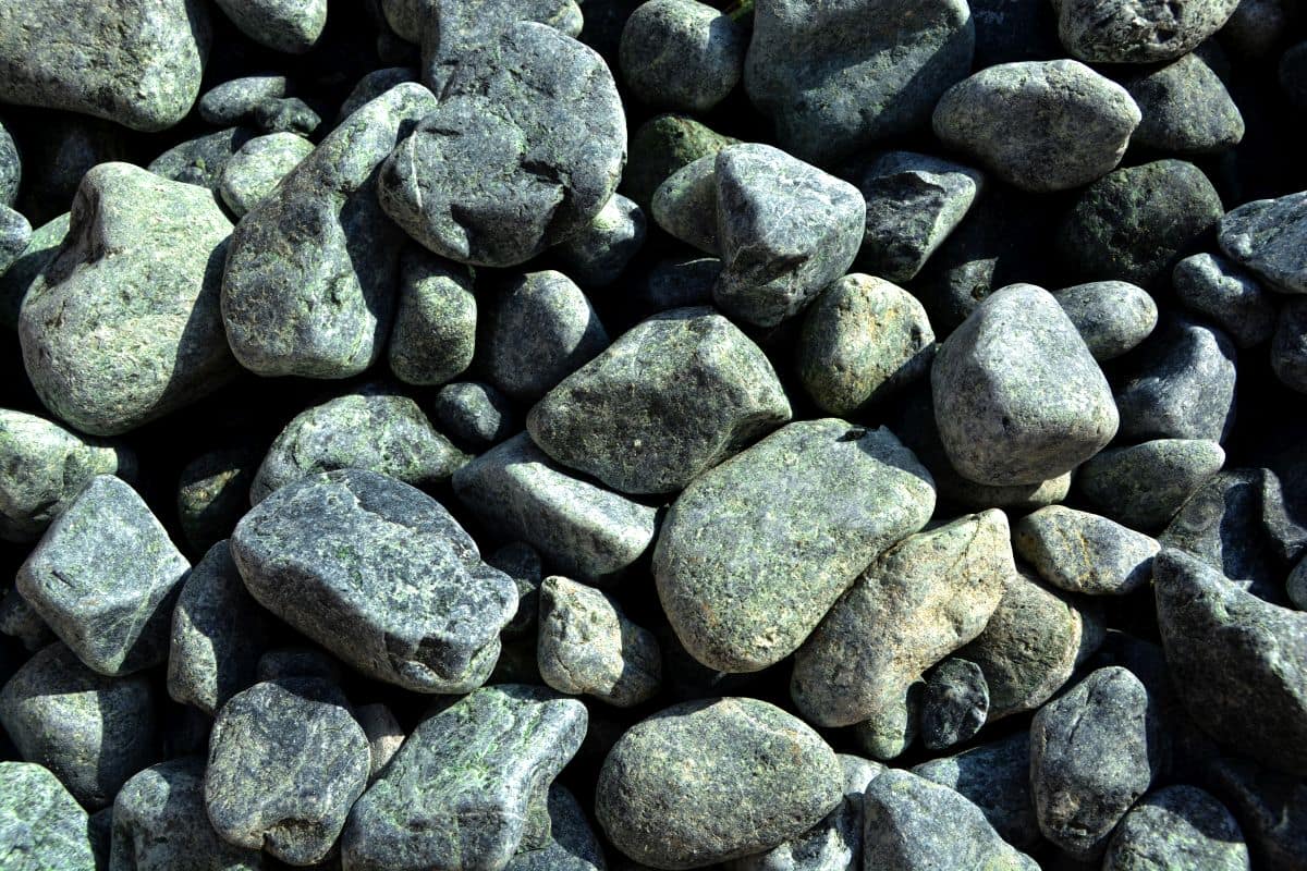Zbliżenie na dużą kolekcję polskich zielonych otoczaków GREEN DREAM S (10/20 mm) ułożonych w nierównym układzie. Gładkie szare i zielone skały różnią się rozmiarem i kształtem, a niektóre mają widoczne tekstury i szczeliny.